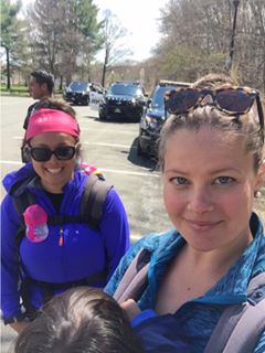 Two women hikers with kids in carriers in a parking lot with emergency vehicles in the background