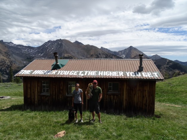 Picture of Pioner cabin with a couple and a dog. Cabin says the higher you get the higher you get